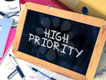 High Priority Handwritten on a Chalkboard. Composition with Small Chalkboard on Background of Working Table with Office Folders, Stationery, Reports. Blurred Background. Toned Image. 3D Render.
