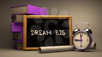 Dream Big - Chalkboard with Hand Drawn Text, Stack of Books, Alarm Clock and Rolls of Paper on Blurred Background. Toned Image.