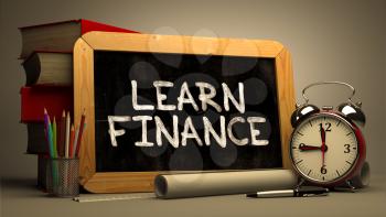 Handwritten Learn Finance on a Chalkboard. Composition with Chalkboard and Stack of Books, Alarm Clock and Rolls of Paper on Blurred Background. Toned Image.