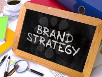 Brand Strategy - Chalkboard with Hand Drawn Text, Stack of Office Folders, Stationery, Reports on Blurred Background. Toned Image.