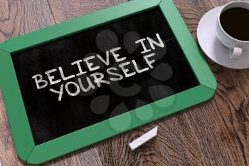 Believe in Yourself - Motivational Quote. Green Chalkboard with Hand Drawn Text and White Cup of Coffee on Wooden Table. Top View.