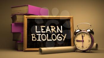 Learn Biology Handwritten by white Chalk on a Blackboard. Composition with Small Chalkboard and Stack of Books, Alarm Clock and Rolls of Paper on Blurred Background. Toned Image.