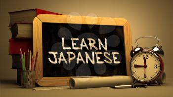 Learn Japanese Handwritten by white Chalk on a Blackboard. Composition with Small Chalkboard and Stack of Books, Alarm Clock and Rolls of Paper on Blurred Background. Toned Image.
