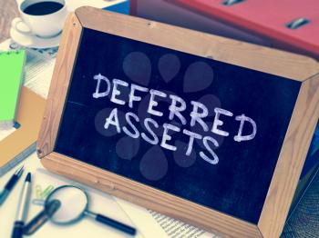 Deferred Assets - Chalkboard with Hand Drawn Text, Stack of Office Folders, Stationery, Reports on Blurred Background. Toned Image.